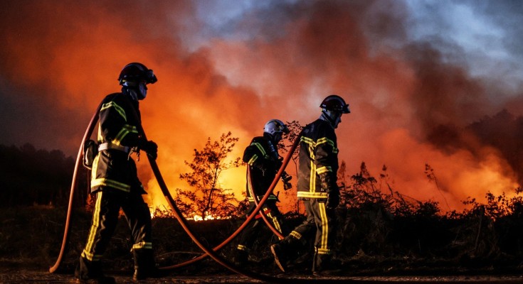 Three firefighters with hoses draped over their shoulders walk across the ground, with smoke billowing behind them.
