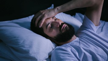 Junger bärtiger Mann liegt im Bett und hält sich die Stirn