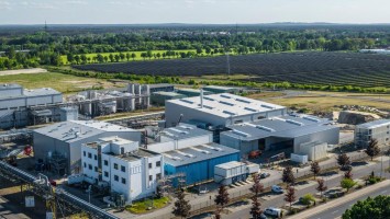 Eine Produktionsanlage, dahinter ein Solarfeld, dahinter Bäume