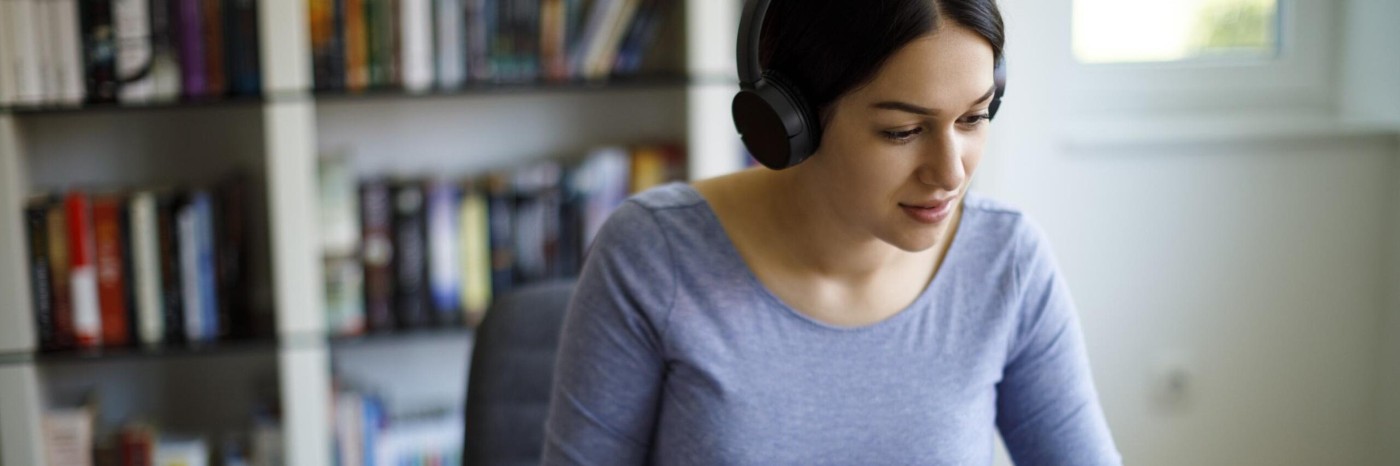 Woman wearing headphones, sat at desktop computer, taking notes on a notepad