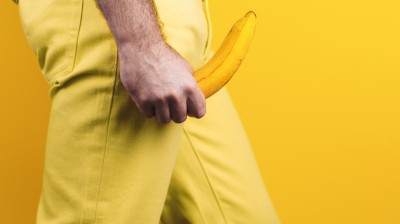 Mann in gelben Jeans hält eine Banane auf Höhe der Genitalien in den Händen