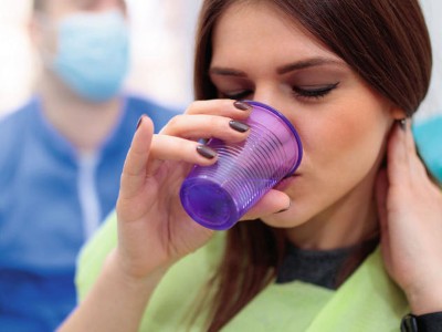 Frau auf zahnärztlichem Behandlungsstuhl trinkt aus einem lila Plastikbecher