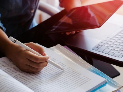 Person mit Stift in der einen Hand über einem geöffneten Buch. In der anderen Hand ist ein Tablet.