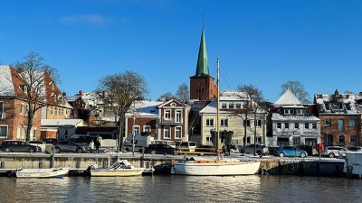 Hafenansicht von neustadt: mehrere kleine Boote liegen vertäut an der Kaimauer. Dahinter sind mehrere Gebäude aus zumeist roten Zieglsteinen sowie eine Kirche mit grünlicher Turmspitze zusehen.