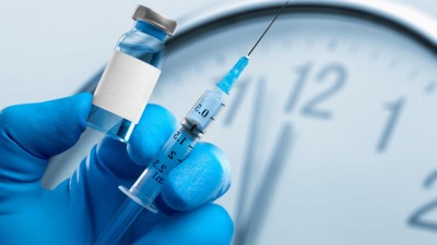 Gloved hand holding a vial and needle in front of a clock