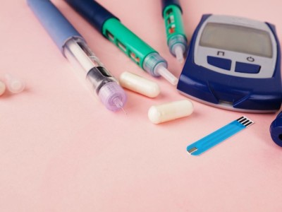 Image is a photograph of insulin pens, capsules, a glucometer and test strips on a pink background