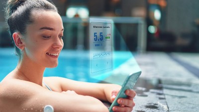 Image is a photograph of a woman monitoring her glucose level with sensor and an app on her phone while training at swimming pool