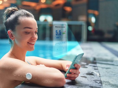 Image is a photograph of a woman monitoring her glucose level with sensor and an app on her phone while training at swimming pool
