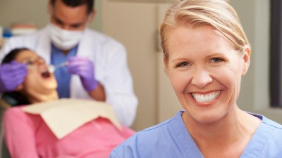 Blonde frau im Vordergrund mit blauem Praxiskittel lacht in die Kamera. Im Hintergrund ein Zahnarzt bei der Behandlung eines Patienten