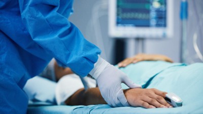 Close-up of a doctor's gloved hand holding the hand of a patient lying in a hospital bed with monitors in the background