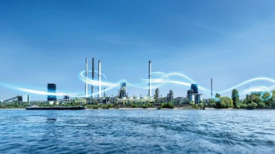Industrielle Anlage, im Vordergrund blau schimmerndes Wasser, im Hintergrund blauer Himmel. 