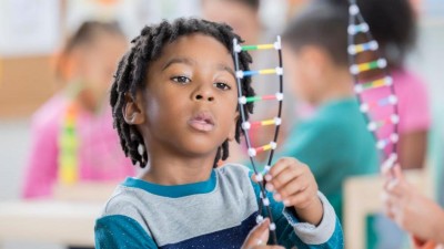 Image is a photograph of an African-American schoolboy in a classroom studying a DNA helix model