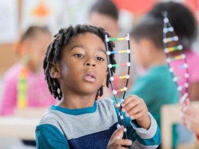 Image is a photograph of an African-American schoolboy in a classroom studying a DNA helix model
