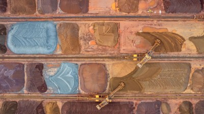 Luftaufnahme eines großen Bereichs mit mehreren farbigen Haufen von Erzrohstoffen, die in rechteckigen Feldern gelagert sind, sowie zwei langen Förderanlagen in der Mitte.