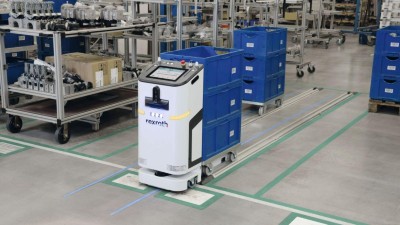 Ein autonomer Transportroboter mit der Aufschrift „Rexroth“ zieht mehrere blaue Kunststoffkisten auf einem Schienensystem durch eine industrielle Produktionshalle.