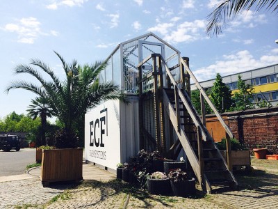 Unten weißer Schiffscontainer, darüber Gewächshaus mit breiter Treppe.