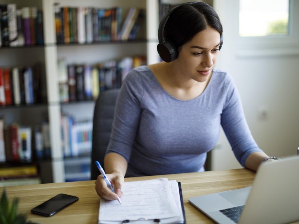 Woman wearing headphones, sat at desktop computer, taking notes on a notepad