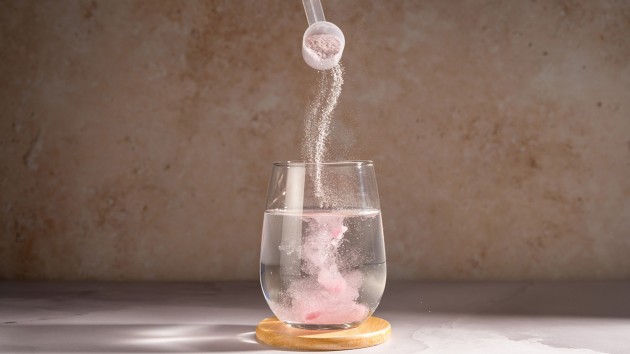 Pink food supplement powder being poured from a scoop into a clear glass of water on a countertop.