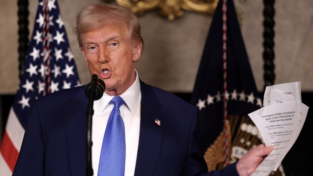 Donald Trump stands at a podium speaking into a microphone, holding up some paperwork.