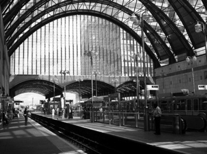 A photograph of a railway station and people waiting on the platform. A train is visible in the last platform.