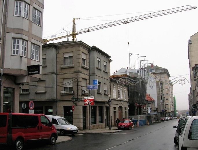 A photograph of a row of single or 2-strey buildings situated on the side of a road, a road junction, along with some vehicles parked by the curb. An arm of a crane is on top.