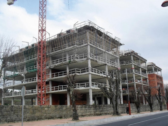 A photograph of a row of 3 buildings, which are under construction, with four floors, surrounded by a boundary and some trees, and situated at the side of a road.