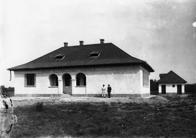 A photograph of a school building named Grof Klebelsberg-Iskola. Two men stands in front of the building. There is a small house with trees in the background.