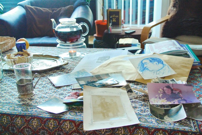 An image of a table covered with printed table cloth. Photographs and documents are placed on the right side of the table while cups, tea pot, tray are kept on the left side of the table. In the background 2 cushioned chairs with a coffee table in between are visible.