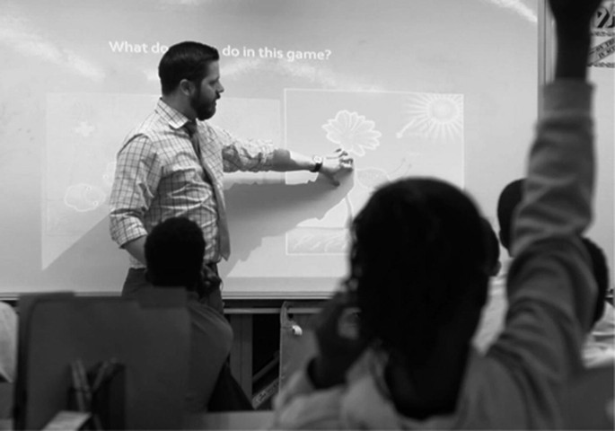 A photo of a classroom displays a man standing and pointing to the slide on the projector. A woman sitting in the foreground raises her hand.