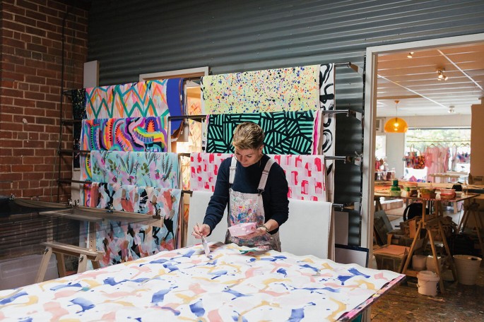 A photograph of a person painting on the fabric spread out on a table. There are various abstract arts of colors and patterns on the background wall.