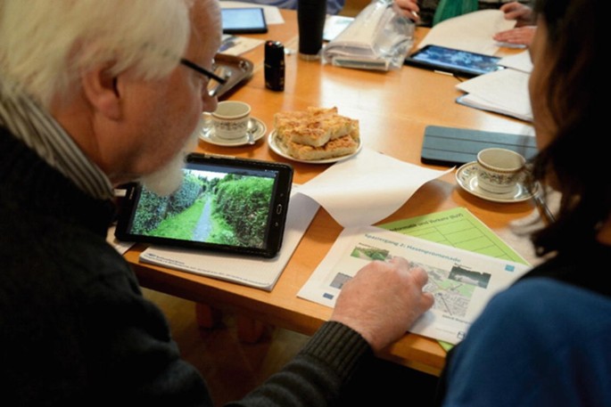 A photo of a man and a woman looking at a route on a map, along with a tablet device and several other objects on a table.