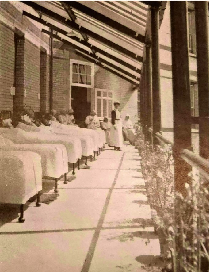 A photograph of a row of patients on beds on a verandah, as the sun shines down on them. A nurse is near the open door.