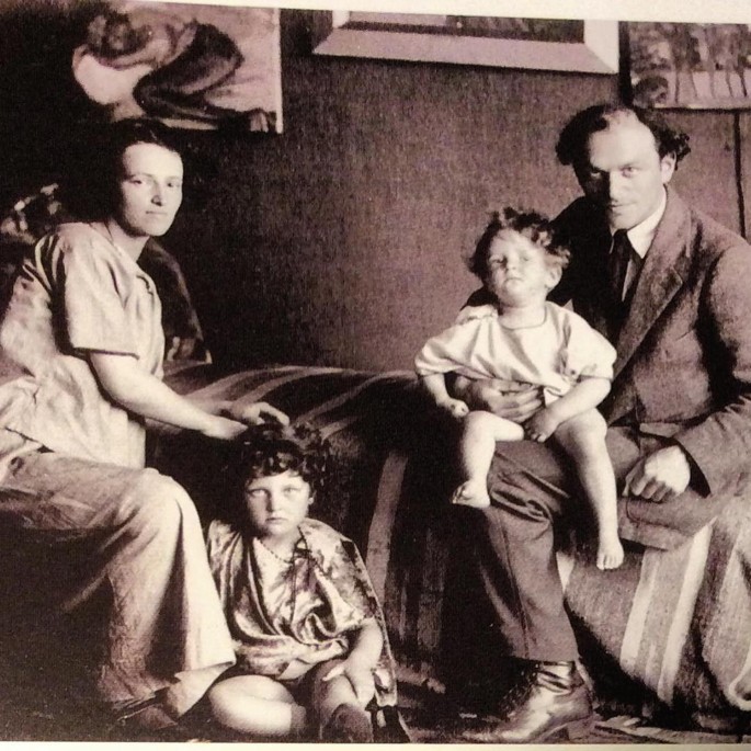 A Winsten family portrait of Clare with her husband and two children. Three paintings on the wall.