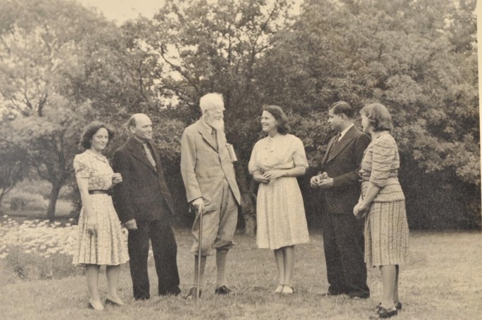 A photo of three couples in formal dress looking at each other. In the background, there are large trees and small plants with flowers.