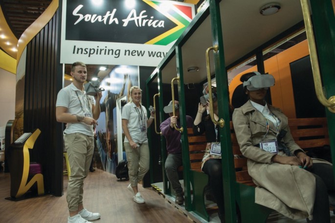 A photograph depicts people seated in rows in an enclosed space wearing visual headsets. A billboard advertisement reads, South Africa, Inspiring new way.