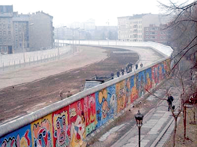 A photo of the Berlin Wall, which has a road between two high walls with several arts in a gradient of colors.