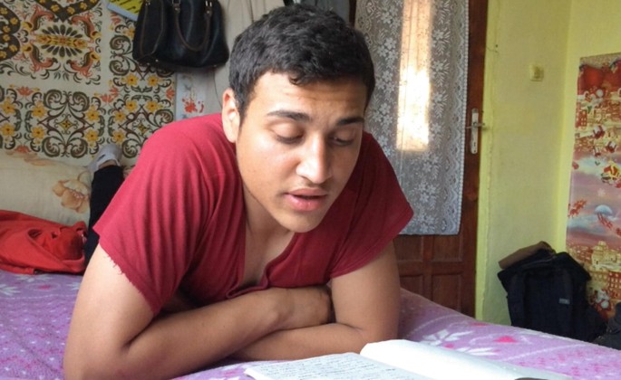 A close-up of a teenage boy lying on his bed. He is in the middle of reading a book that is on the bed in his front.
