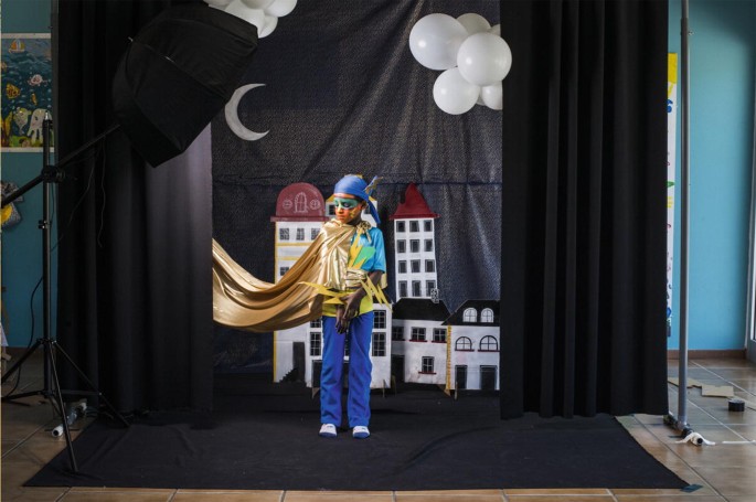 A portrait of a young boy in a photo studio. He is dressed up like a fictional superhero. The background is decorated with cardboard houses and balloons.