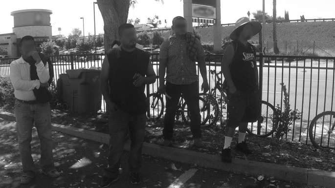 A grayscale view of a group of men standing near the fence on a sidewalk. Their faces are blurred. Cycles are parked beside them.