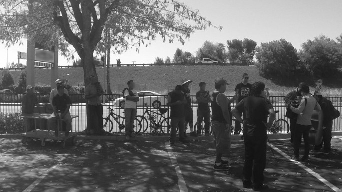 A grayscale view of a group of men standing in front of a fence on a sidewalk. A few men on the right side are playing different musical instruments. The others look at them.