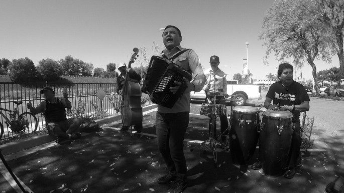 A grayscale view of a band. A man sings and plays the accordion in front. The other men behind him play drums, violins, and other musical instruments.