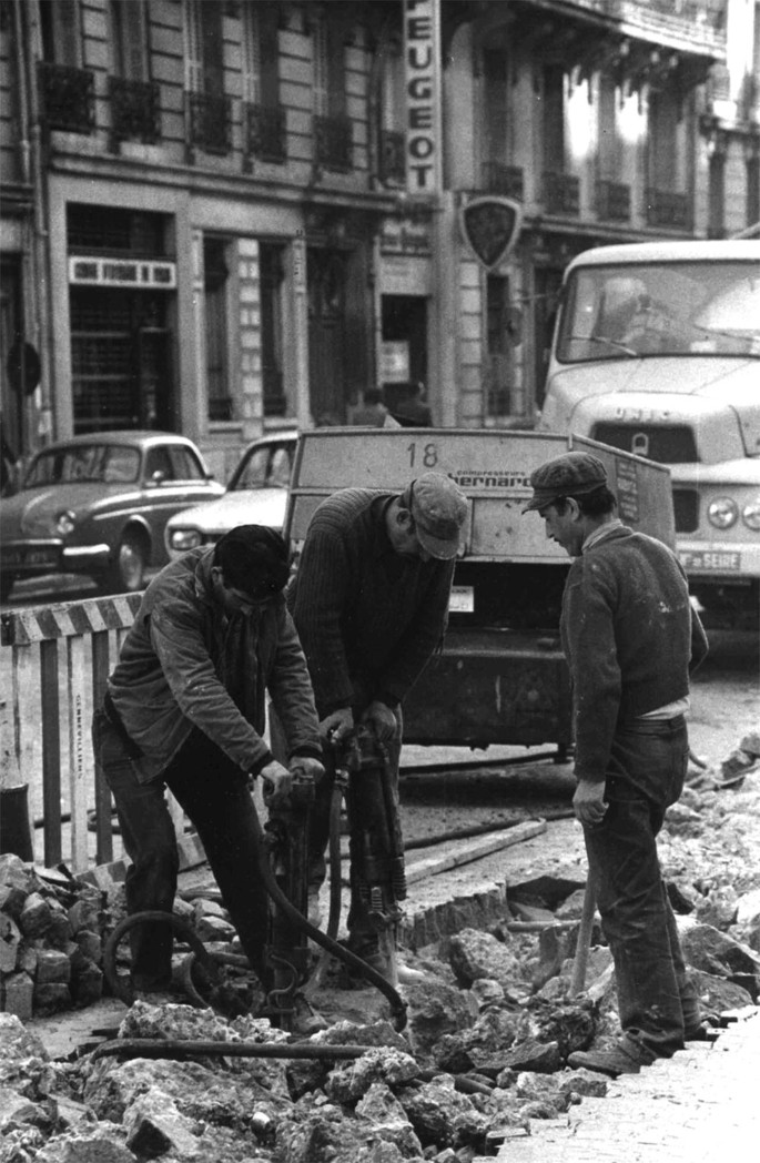 A grayscale view of two men digging up the sidewalk of a street. Another man stands in front of them with a shovel in his hand. The background has cars parked on the road.