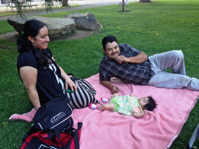 A man and a woman look at the baby in front of them and smile. The baby is lying on a cloth on the grass. The background has a grassy field.