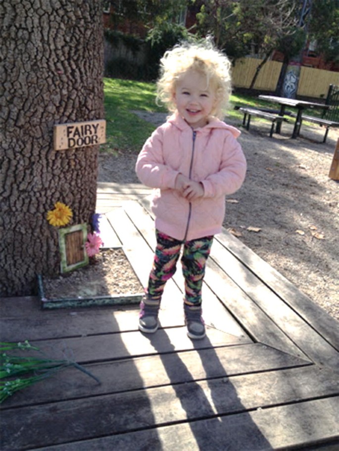 A photograph of a kid standing near a tree to which a fairy door card is attached.