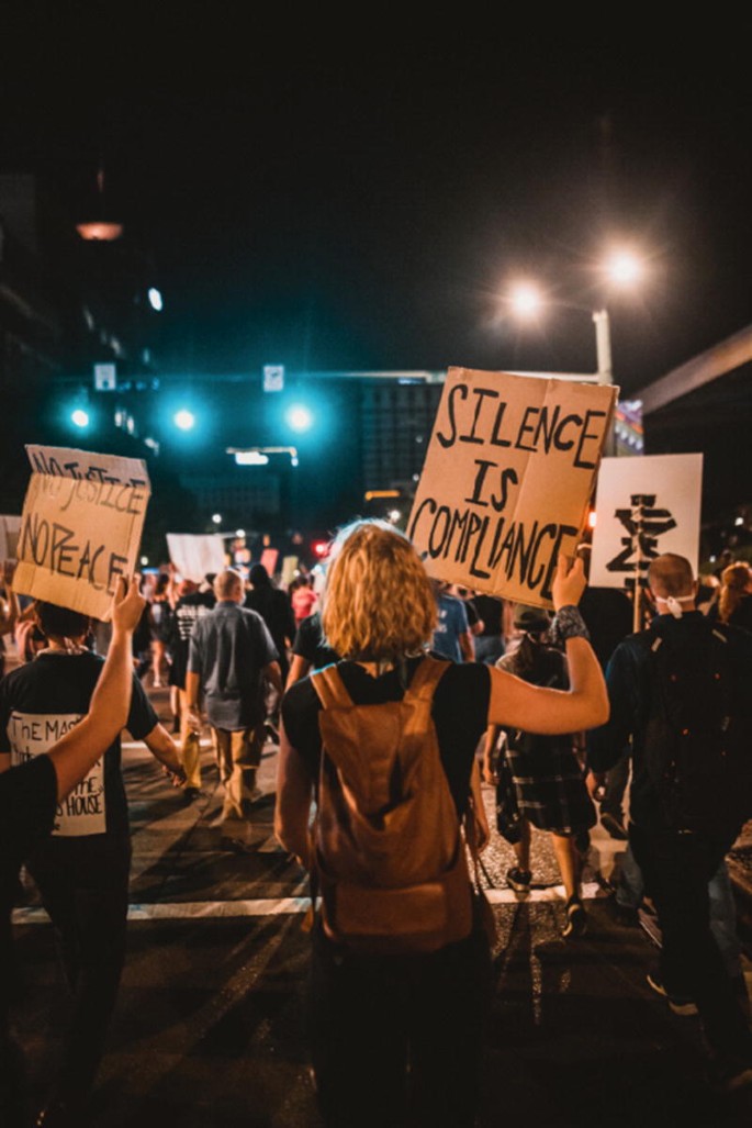 A back view of a group of men and women holding boards that read "silence is compliance."