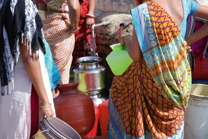 A photograph of a cropped view of a group of women lining up in front of two knobs. The women carry pots and buckets in their hands.
