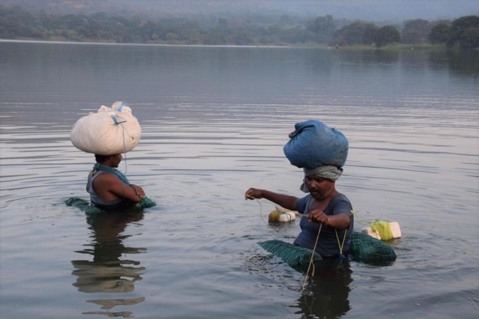 A landscape view of two men in the water. Both of them have sacks on their heads. The right man stretches a rope with his hand.