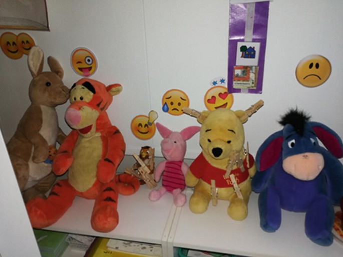 A front view of several soft toys lined up on a desk. The wall behind them is pasted with emoji stickers.