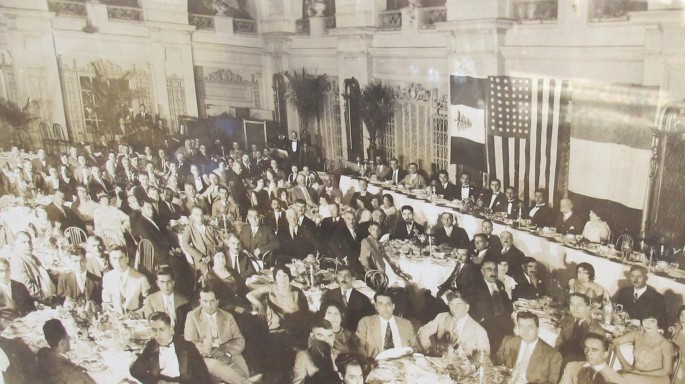 A photo of a crowd of people gathering, organizing themselves into rows, and sitting at round tables. The tables are adorned with food arrangements.