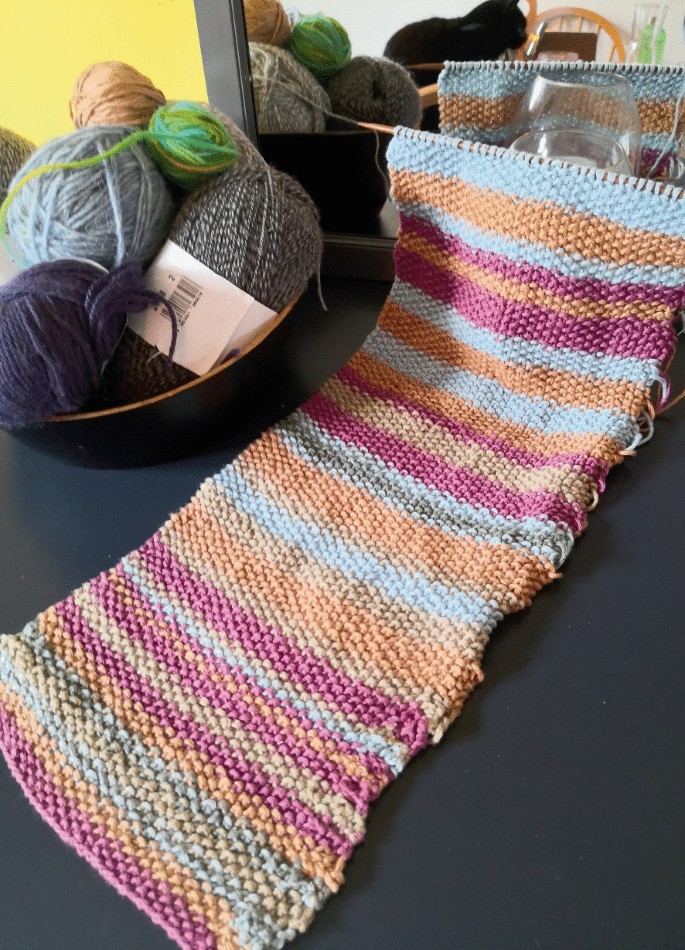 A photograph of a knitted scarf with a knitting needle on a table in front of a mirror. A basket on the left has 5 balls of wool.